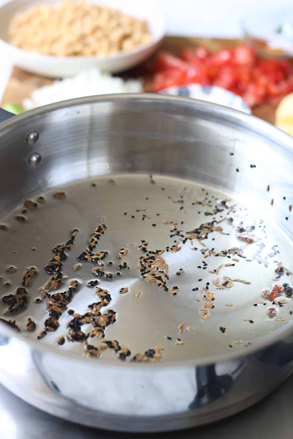 A close-up of a stainless steel pan with toasted cumin seeds, with blurred chopped onions, tomatoes, and chickpeas on a cutting board in the background-perfect for preparing flavorful aloo chana.