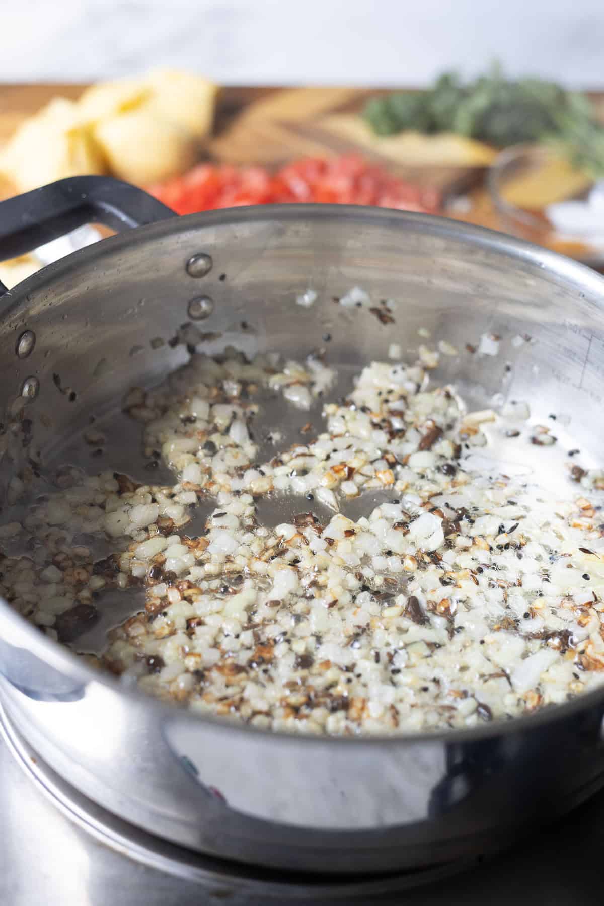 Diced onions and minced garlic are being sautéed in a stainless steel pan, with a blurred background showing chopped vegetables and herbs on a wooden cutting board for an Aloo chana preparation.