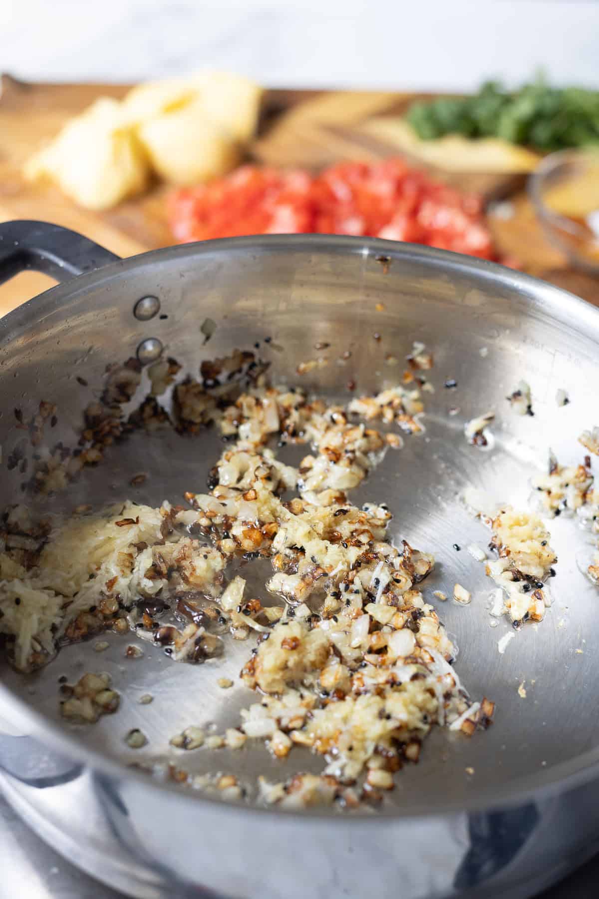 Finely chopped onions and garlic are sautéing and browning in a stainless steel pan, creating a flavorful base for aloo chana. In the background, chopped tomatoes, herbs, and pineapple pieces rest on a wooden cutting board.