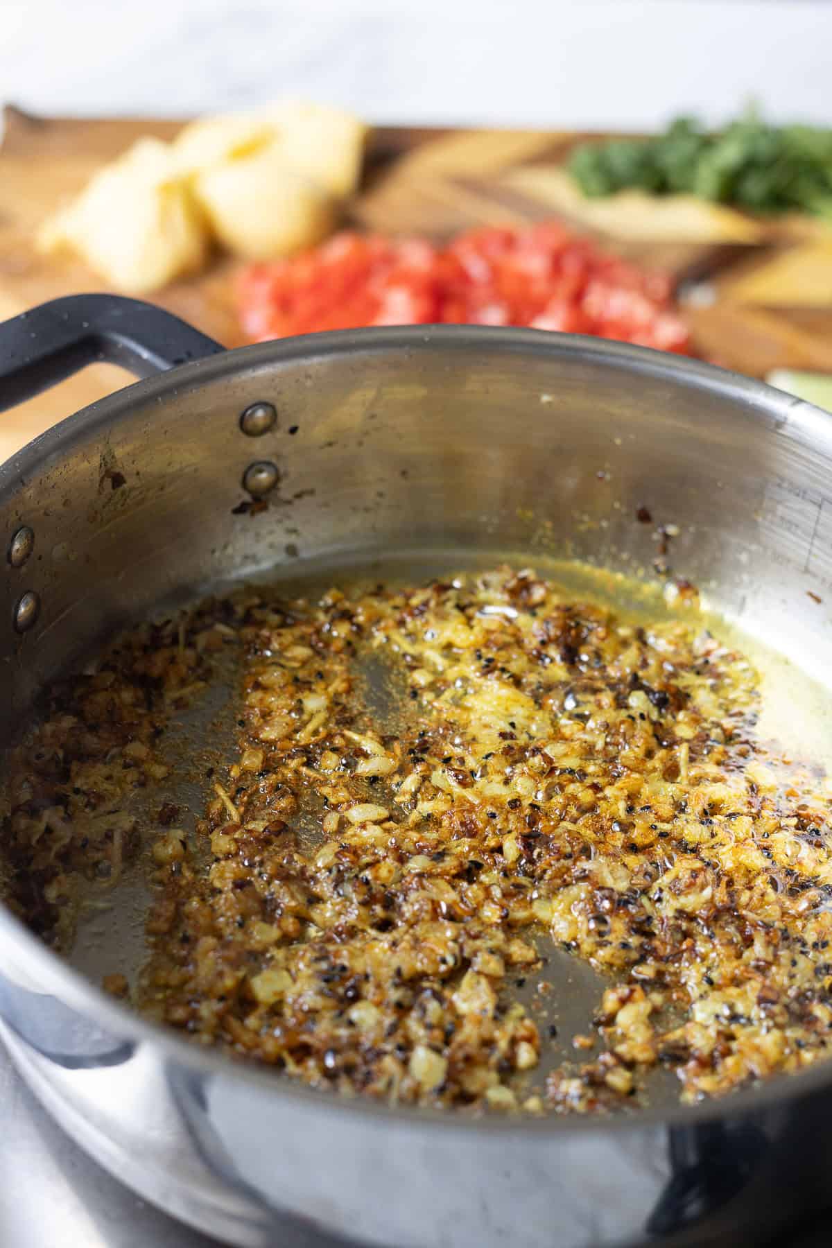 A close-up of onions sautéing and browning in a metal saucepan on a stove, with ingredients for aloo chana-chopped tomatoes, herbs, and ginger-on a wooden cutting board in the blurred background.