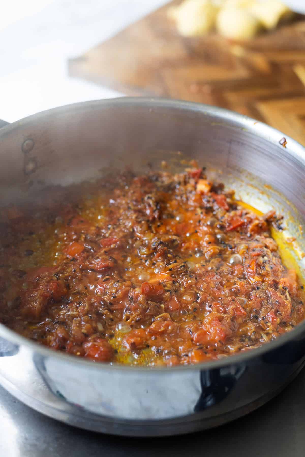 A metal pan on a stove contains a simmering tomato-based sauce with spices, possibly for aloo chana. In the blurred background, there is a wooden cutting board with dough balls on it.
