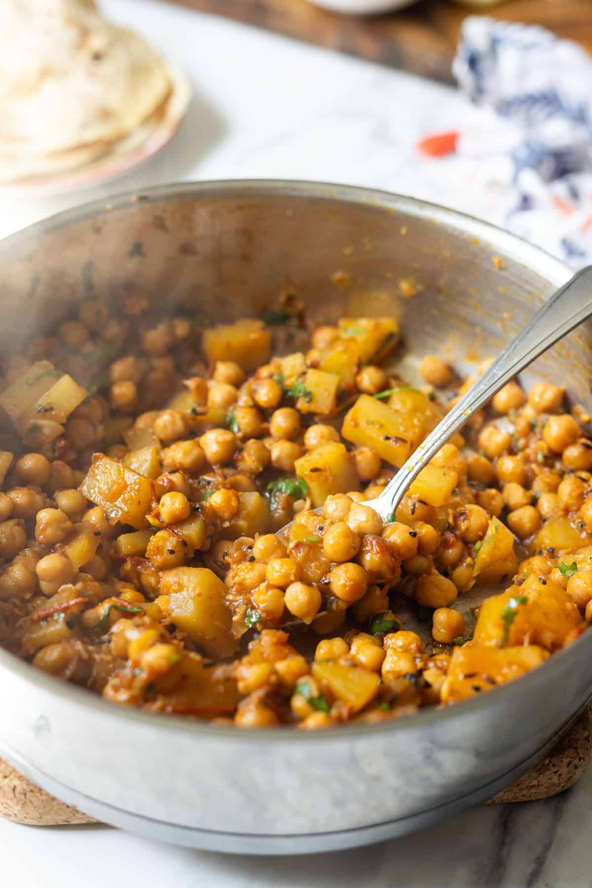 A close-up of a metal pan filled with aloo chana, a chickpea and potato curry garnished with herbs, with a spoon resting inside. In the background, there are blurred flatbreads on a plate.