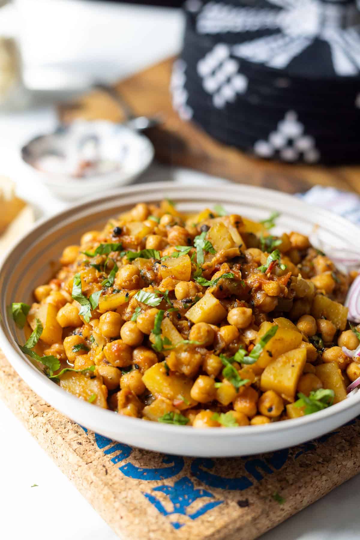 A close-up of a bowl filled with Aloo chana—cooked chickpeas and diced potatoes—garnished with fresh chopped herbs, sitting on a cork mat. Sliced red onion and a patterned dish are visible in the background