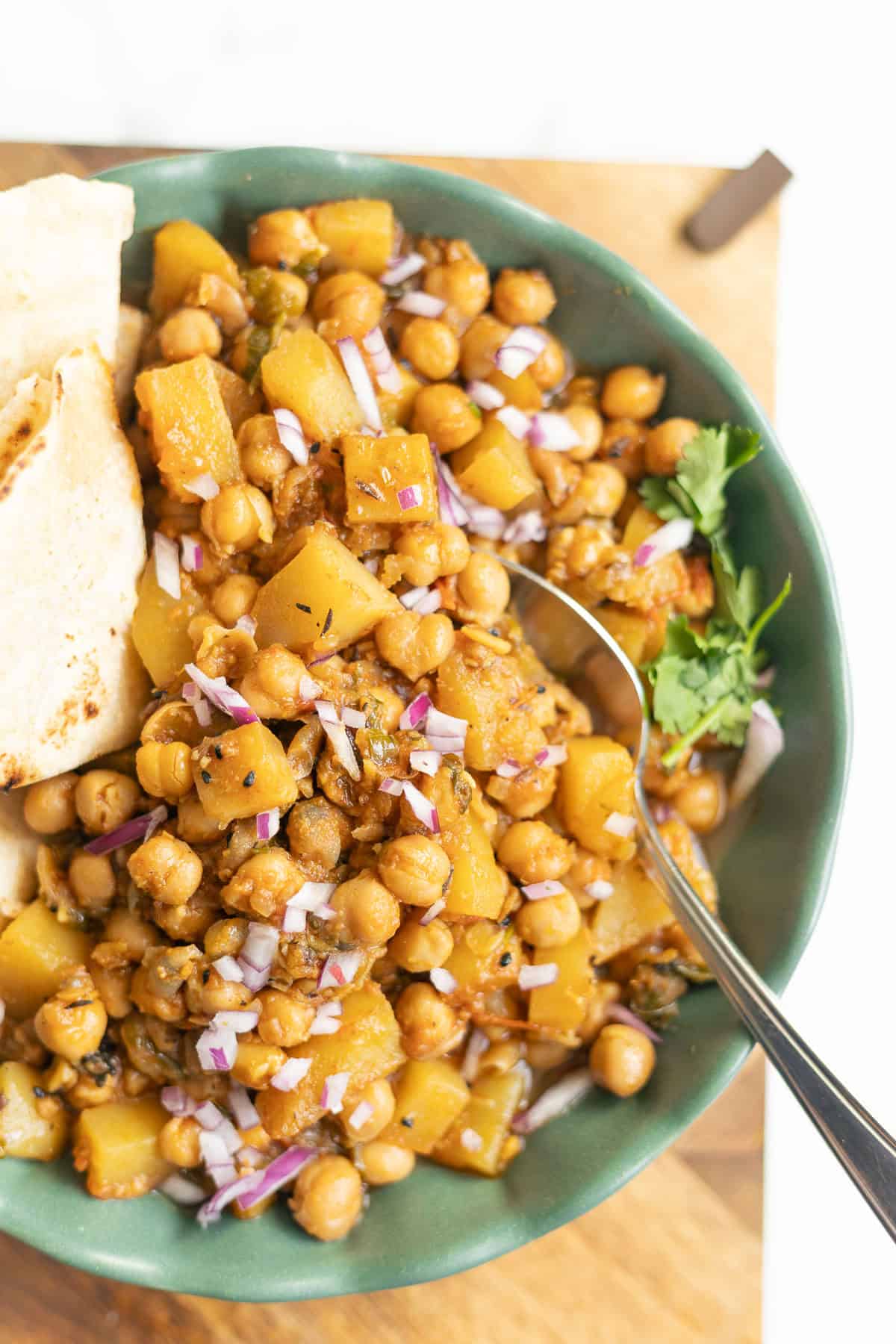 A green bowl filled with aloo chana, a flavorful chickpea and potato curry, garnished with chopped red onions and cilantro. Flatbread pieces are on the side, and a spoon rests in the bowl on a wooden surface.