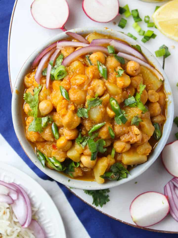A bowl of aloo chana curry garnished with chopped onions, green chilies, and cilantro, surrounded by sliced radishes, green onions, lemon wedge, and a plate of rice on the side.