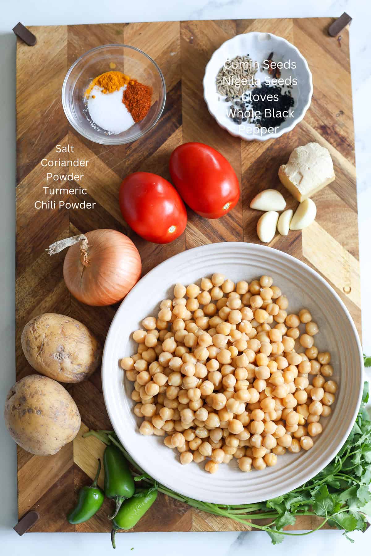 A wooden board holds fresh ingredients for Aloo chana: chickpeas, tomatoes, onion, potatoes, green chilies, cilantro, ginger, garlic, and bowls of spices-salt, coriander powder, turmeric, chili powder, cloves, nigella seeds, cumin seeds, and black pepper.