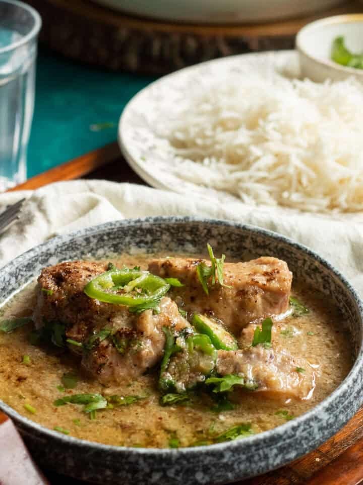 A bowl of creamy white chicken curry garnished with sliced green chilies and cilantro, served alongside a plate of steamed white rice. A glass of water is also visible in the background.