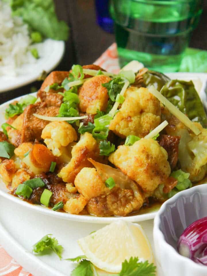 A bowl of Indian-style cauliflower and potato curry inspired by Pakistani cuisine, garnished with cilantro and surrounded by lemon wedges, green onions, chilies, red onion, and rice. A wooden spoon rests beside the bowl.