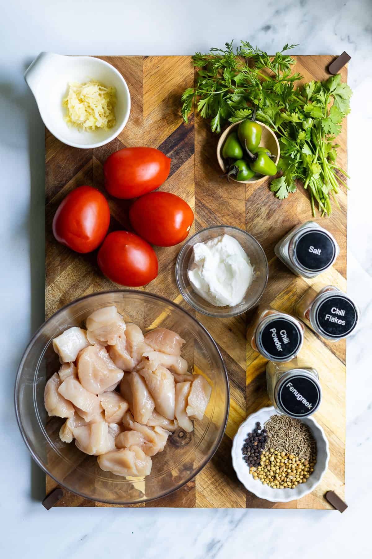A wooden board holds raw diced chicken for Karahi Chicken, whole tomatoes, minced garlic, yogurt, fresh cilantro, green chilies, and small bowls of salt, chili flakes, coriander seeds, black pepper, and cumin seeds.