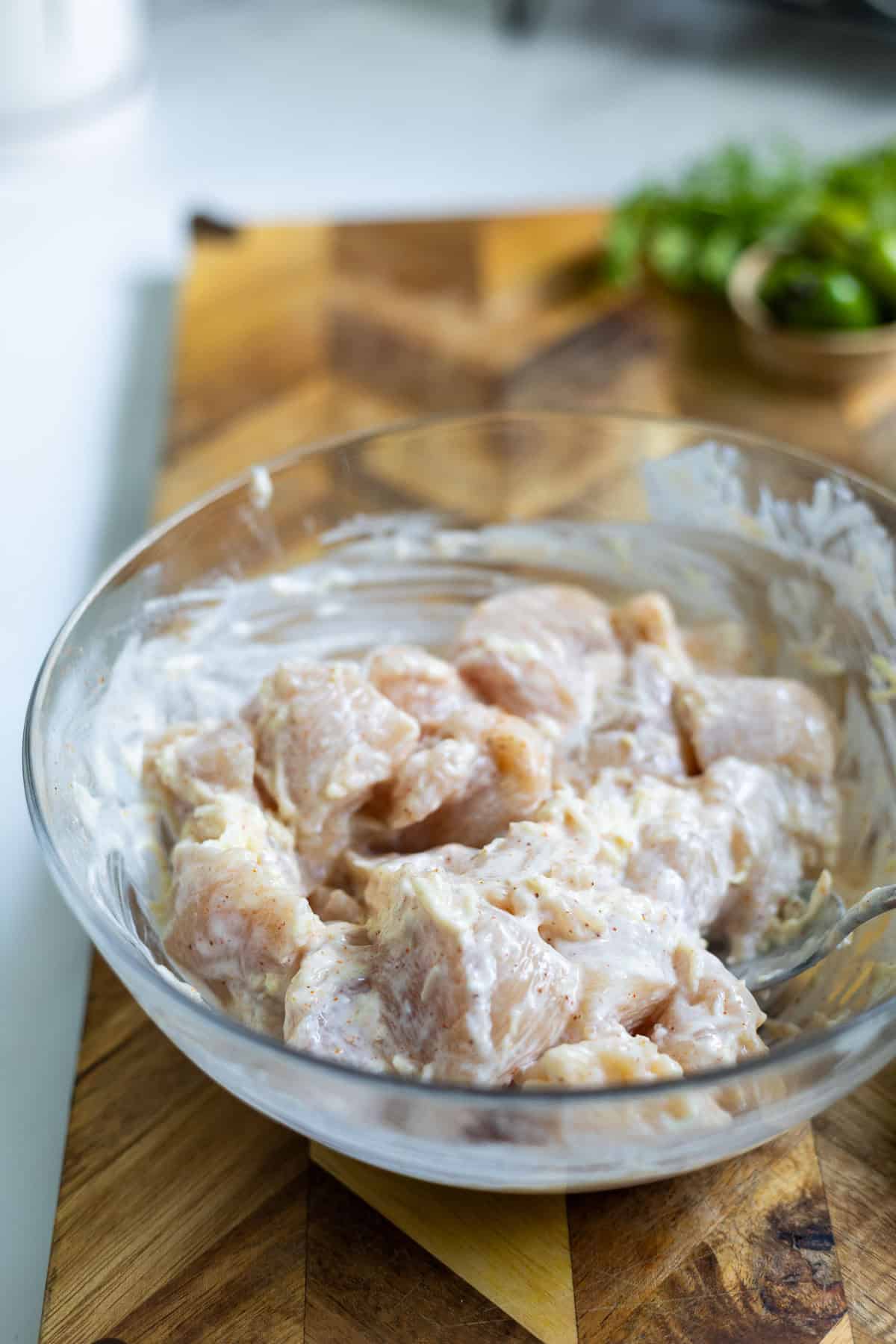 Raw chicken pieces for Karahi Chicken are coated in a creamy marinade, sitting in a glass bowl on a wooden cutting board, with fresh herbs blurred in the background.