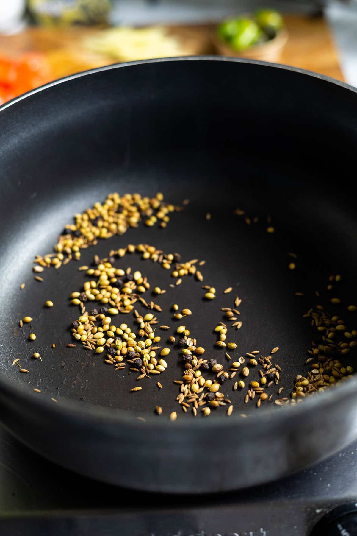 Whole spices, like coriander and cumin seeds, are being dry roasted in a black nonstick pan on the stovetop, creating the aromatic base often used in dishes such as Karahi Chicken. The background blurs with kitchen ingredients.