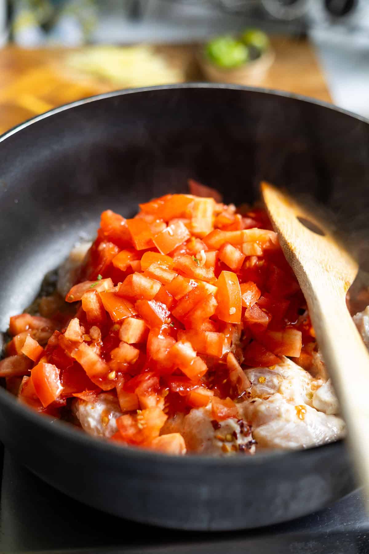 Chopped tomatoes simmer with pieces of chicken in a black frying pan, as a wooden spoon stirs the flavorful Karahi Chicken mixture. Steam rises gently, set against a blurred kitchen background.