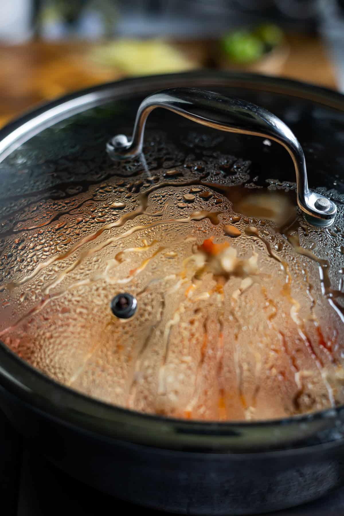 A close-up of a pot with a glass lid covered in condensation, showing steam and droplets inside. Karahi Chicken with visible bits of vegetables can be seen through the lid. The background is slightly blurred.