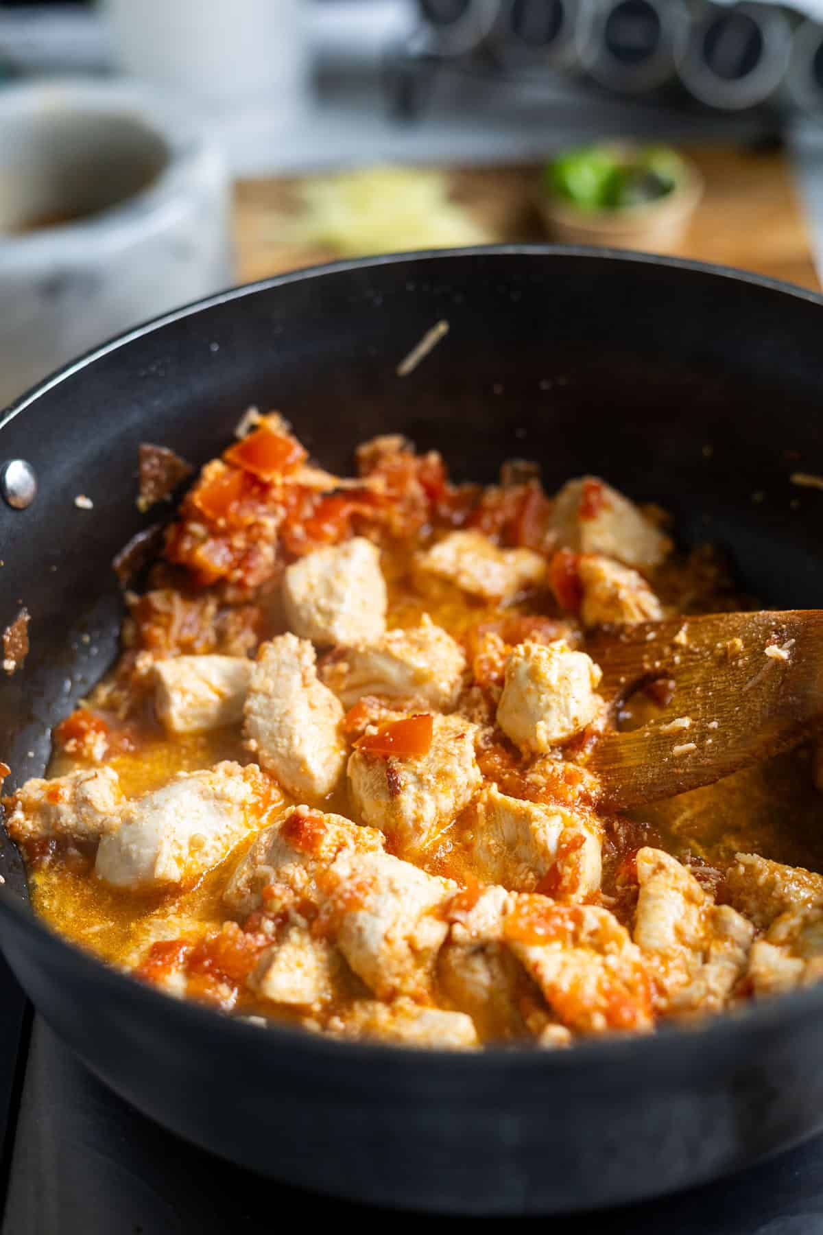 Chunks of chicken simmering in a skillet with chopped tomatoes and sauce, being stirred with a wooden spoon-just like an authentic Karahi Chicken. Kitchen utensils and ingredients are blurred in the background.