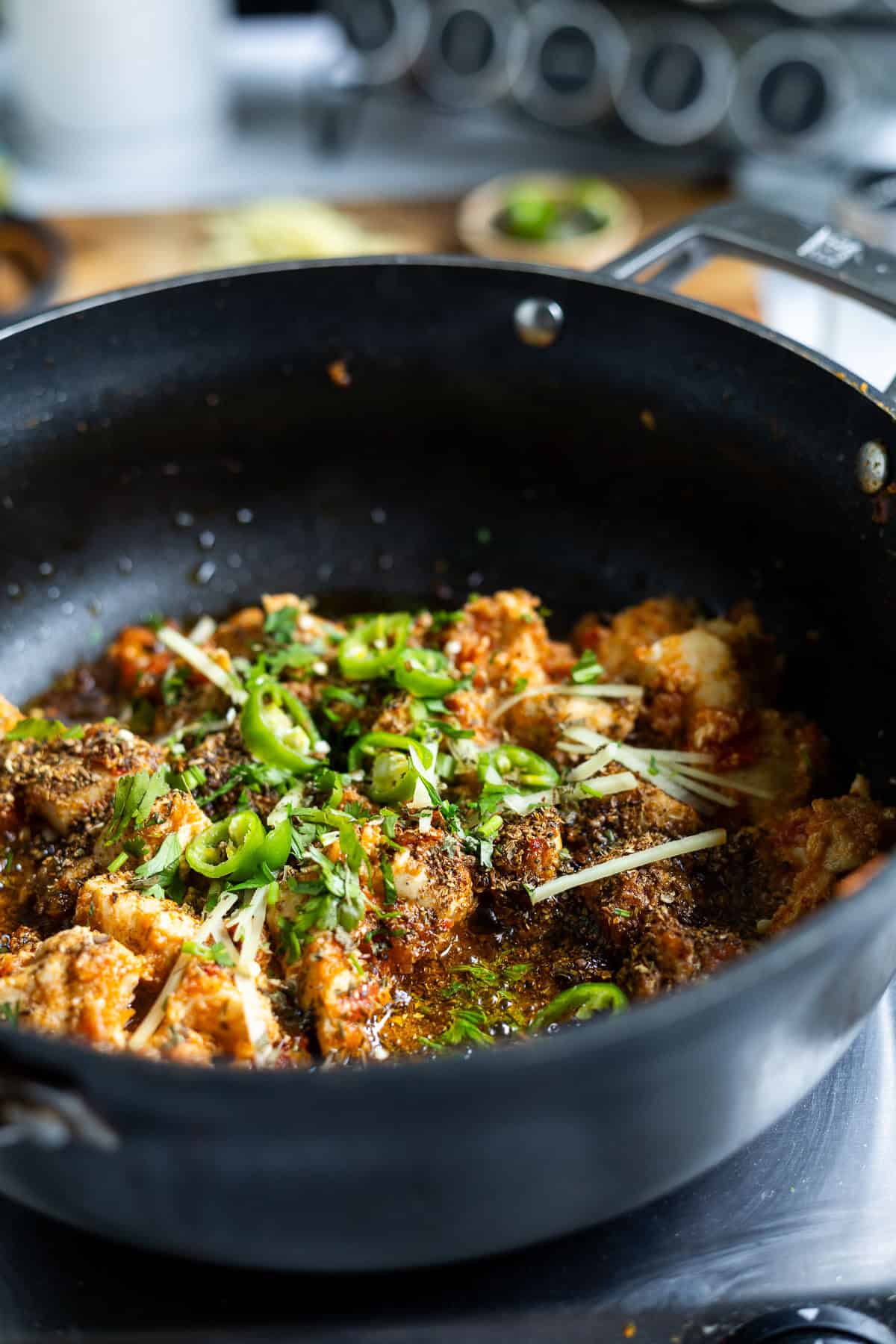 A close-up of a black pan filled with spiced Karahi Chicken curry, garnished with sliced green chilies, ginger strips, and chopped cilantro, cooking on a stove with kitchen utensils blurred in the background.