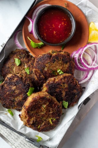 Five Shami Kabab spiced meat patties garnished with herbs, served on parchment paper with sliced red onions, lemon wedges, and a small bowl of red dipping sauce on a brown plate.