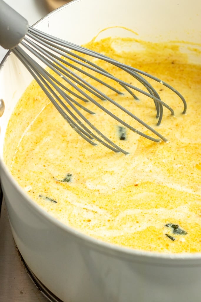 A metal whisk stirs a creamy yellow Kadhi Pakora soup with green herb pieces in a white pot.